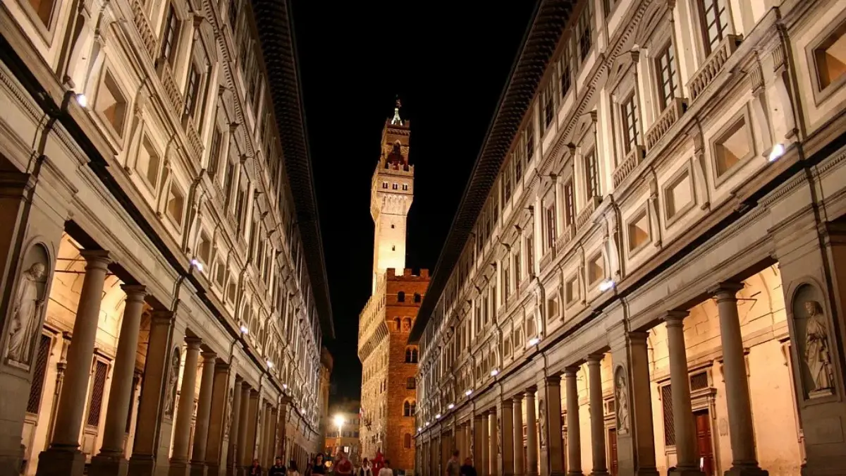 Night view of the Uffizi Gallery in Florence with people walking and the Palazzo Vecchio tower in the background.